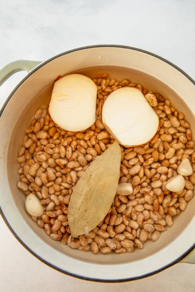 Pot of beans, onions, garlic cloves, and a bay leaf submerged in water.