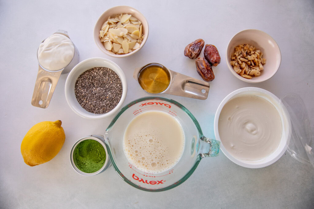 Work surface covered with yogurt, cream cheese, chia seeds, agave syrup, almonds, dates, matcha, lemon, milk, and banana chips.