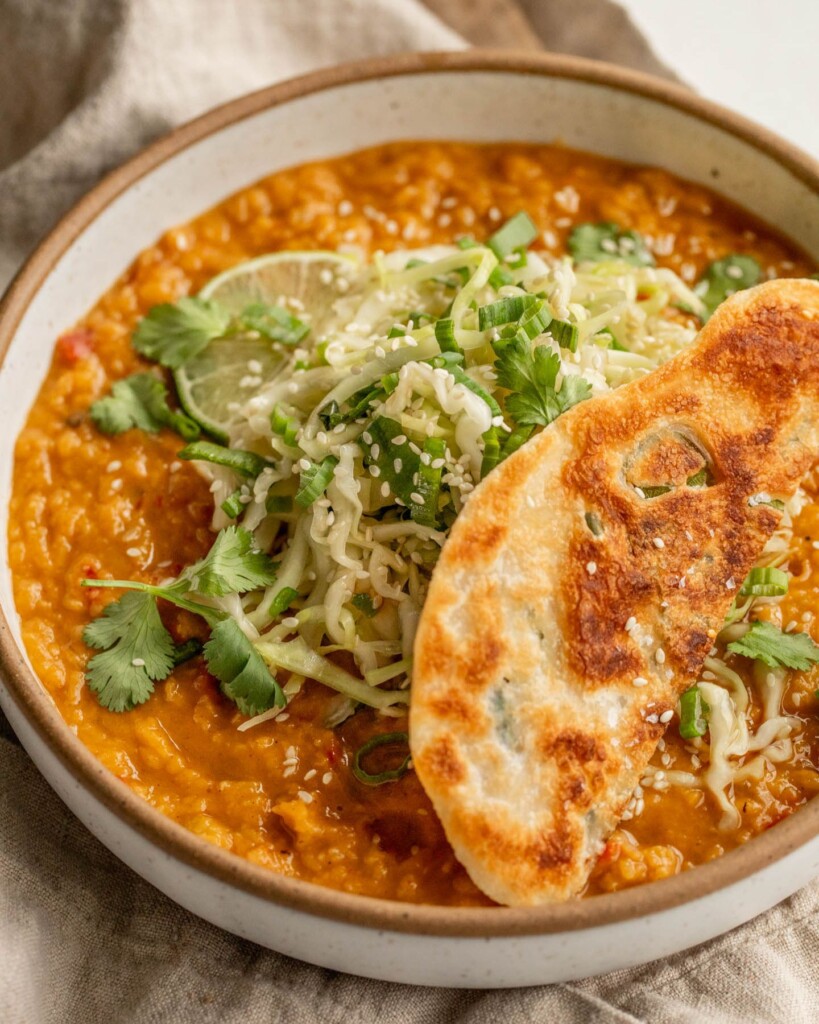 Close up of a bowl of creamy red lentils topped with cabbage slaw and a piece of a crispy scallion pancake on top.