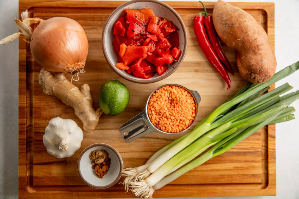 Cutting board topped with a sweet potato, bowl of diced roasted bell peppers, red lentils, scallions, spices, lime, onion, ginger, bulb of garlic, and chillies.