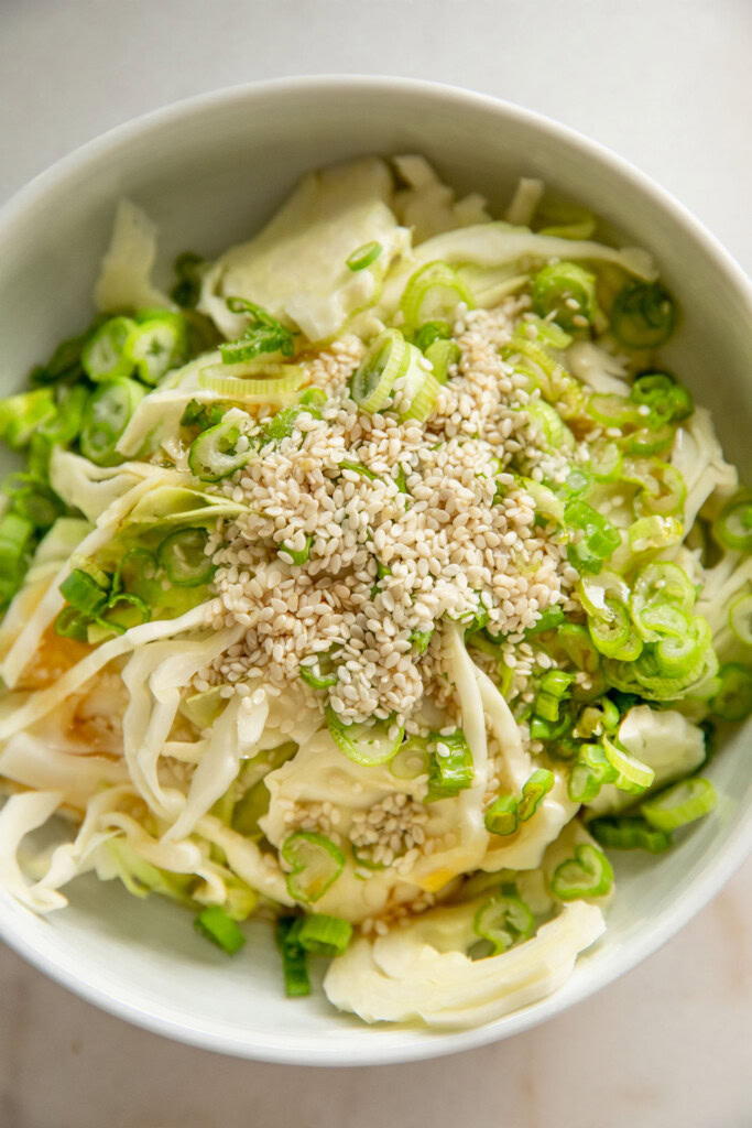 Bowl of shredded cabbage with sesame seeds, vinegar, oil, and scallions.