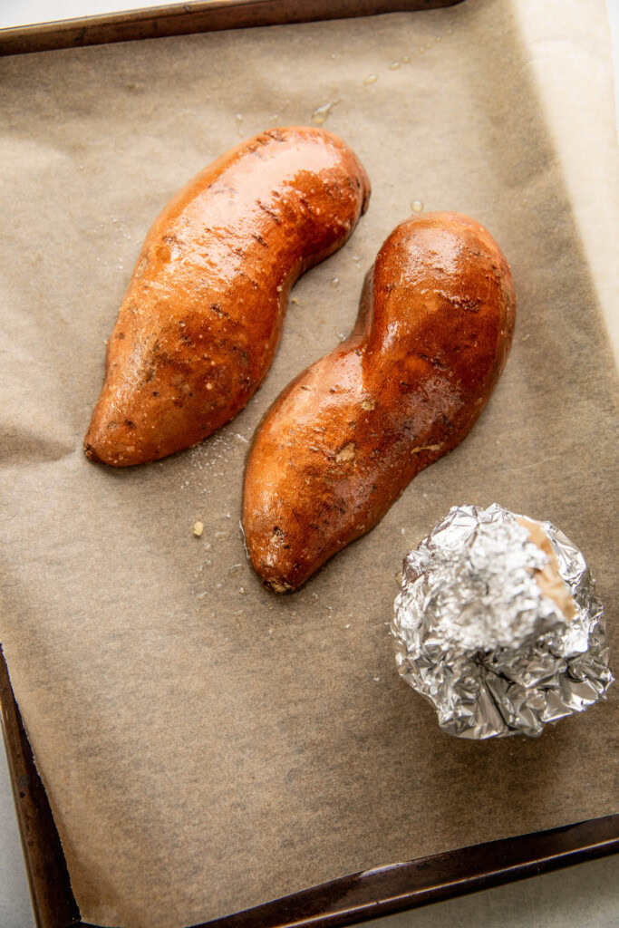 Garlic bulb wrapped with foil and halved sweet potatoes on a baking tray.