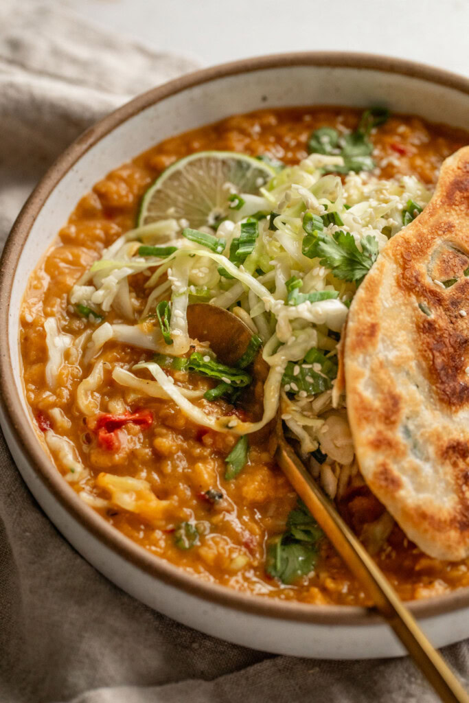Close up of a bowl of lentils being scooped up with a spoon and topped with cabbage slaw and a crispy piece of a scallion pancake on top.