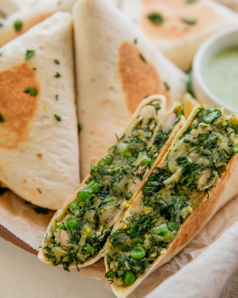 A potato leek wrap cut in half on a plate filled with toasted wraps behind it and a bowl of herby yogurt sauce.