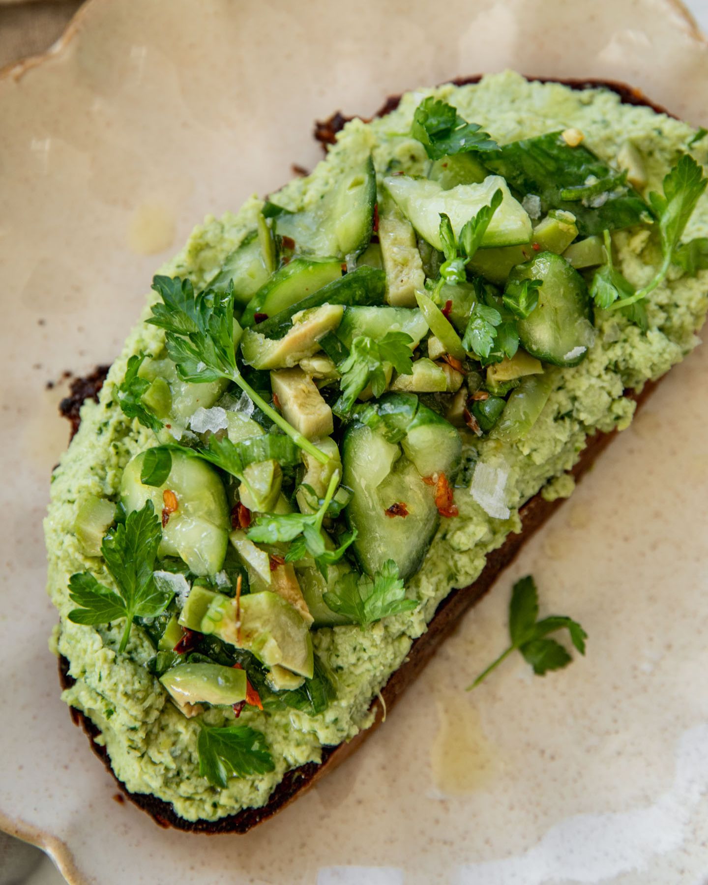 Close up of a toast on a plate layered with artichoke edamame spread and topped with a marinated cucumber salad.