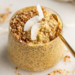 A cup filled with mango chia pudding that is topped with some crumble and coconut flakes, being dug into with a spoon.