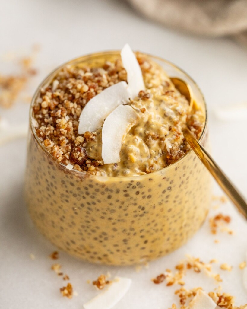 A cup filled with mango chia pudding that is topped with some crumble and coconut flakes, being dug into with a spoon.