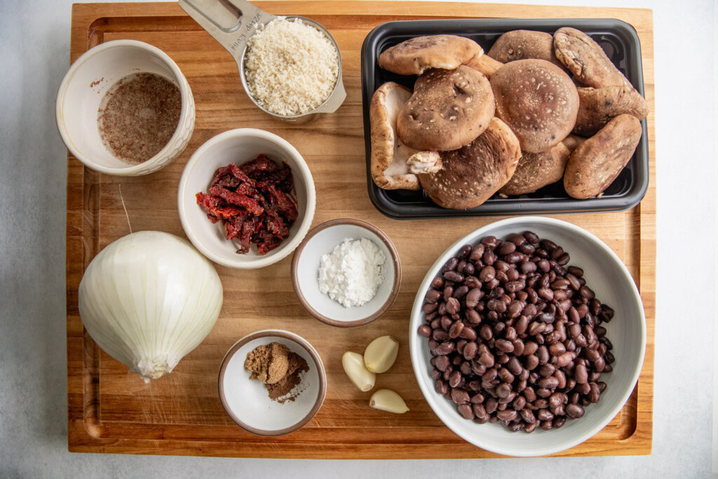 Cutting board with mushrooms, black beans, garlic, cornstarch, spices, sun-dried tomatoes, onion, flax egg, and breadcrumbs.