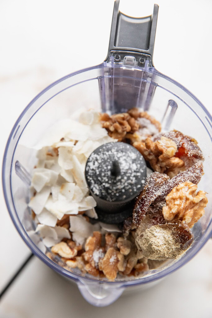 Food processor filled with dates, coconut flakes, cardamom, and walnuts.