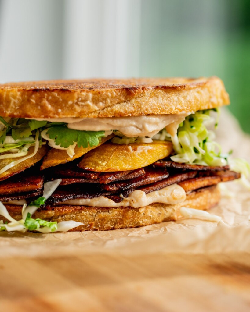 A sandwich layered with white bean spread, smoky chipotle tofu slices, baked plantains, and cabbage slaw.