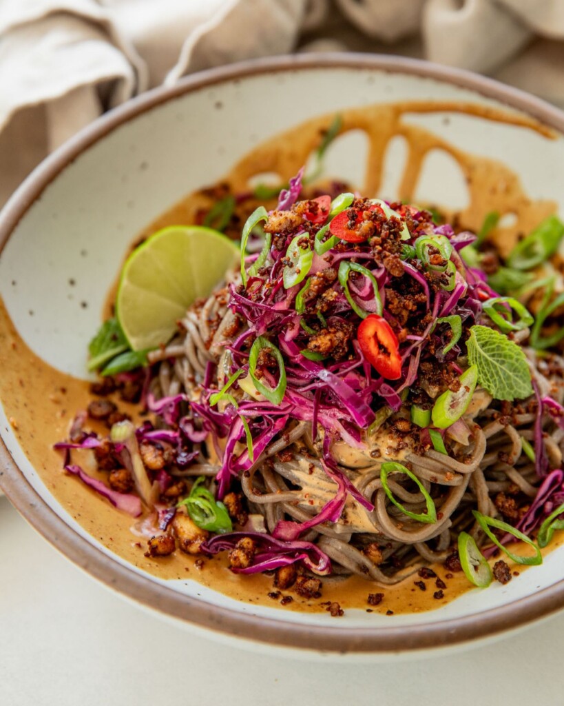 Bowl ladled with sesame sauce and layered with soba noodles, cabbage slaw, crumbled tofu, and scallions.