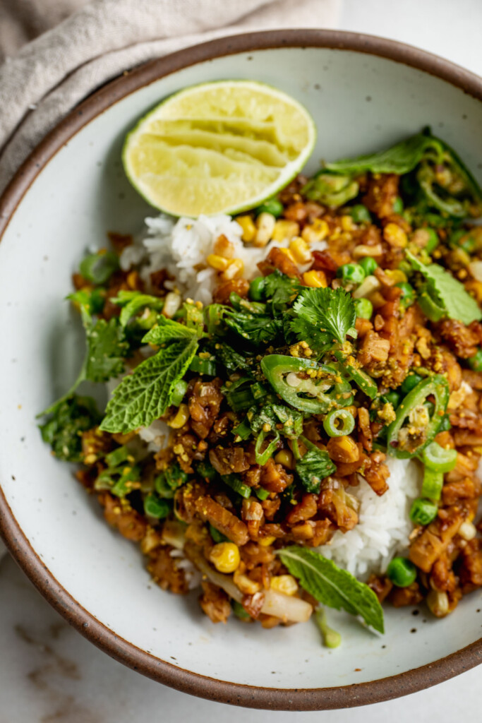 Close up of a bowl of rice topped with sticky tempeh and scallion salad with a wedge of lime on the side.