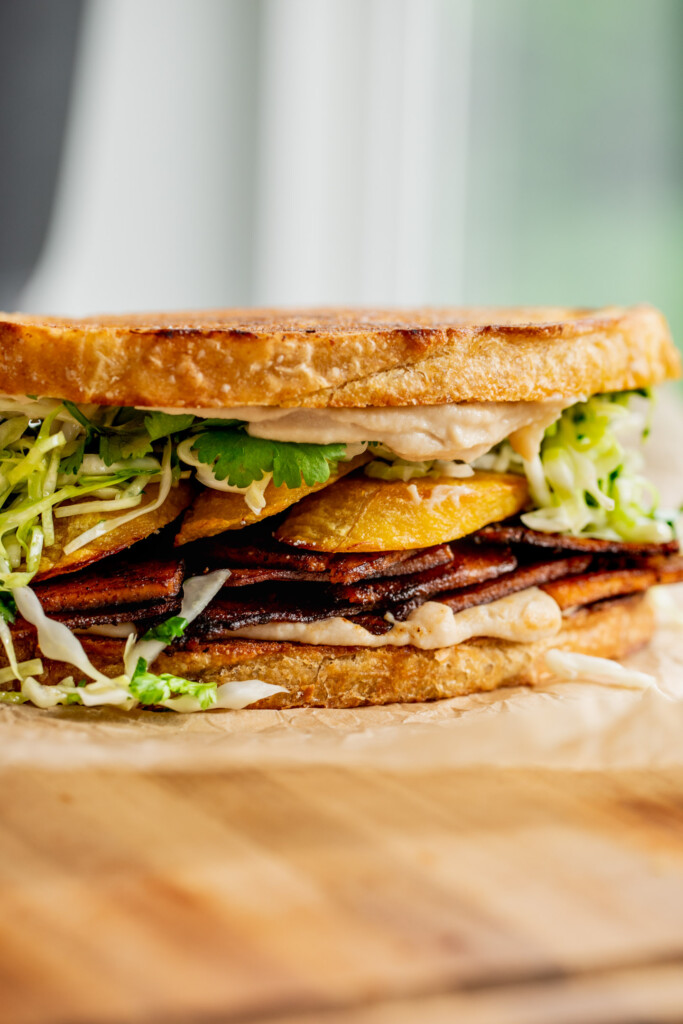 A sandwich layered with smoky tofu deli slices, baked plantain, and cabbage slaw.