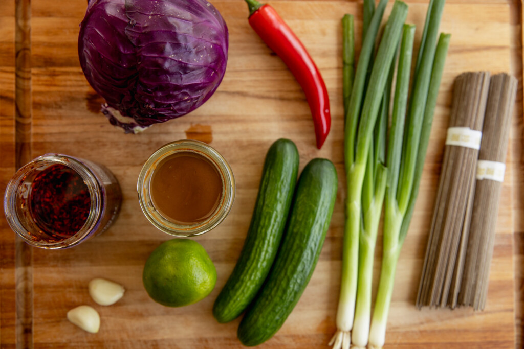 Cutting board with red cabbage, cucumbers, scallions, red pepper, lime, sesame paste, chili crisp oil, and garlic cloves.