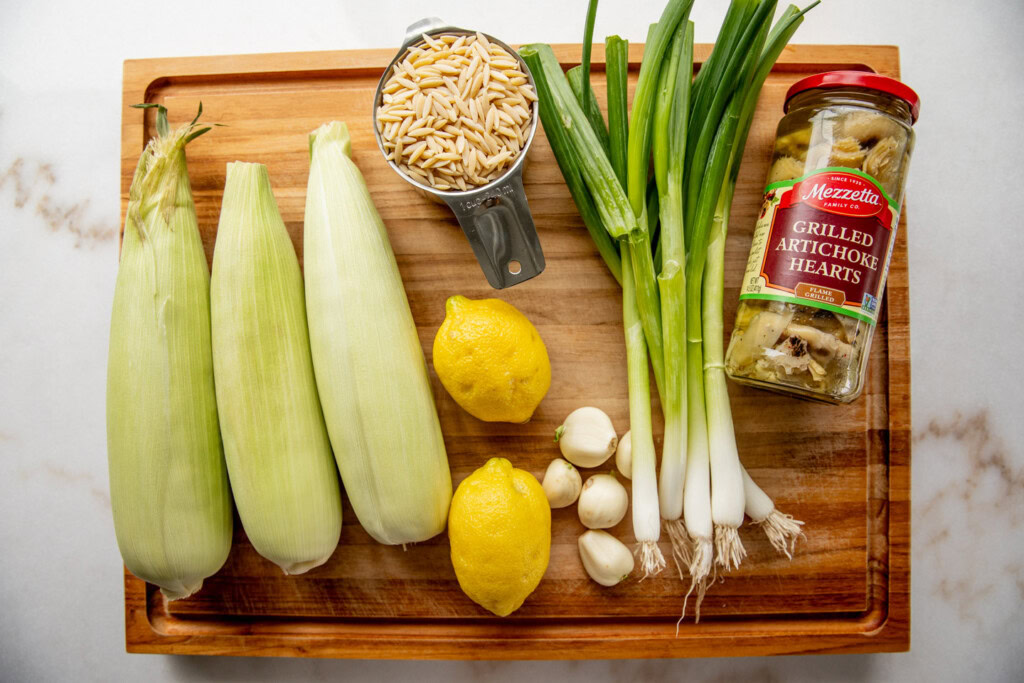 Corn cobs, lemons, garlic, scallions, artichoke hearts, and orzo on a cutting board.