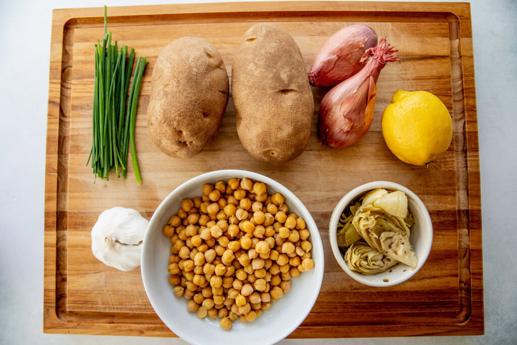 Cutting board with chives, chickpeas, artichoke hearts, lemon, shallots, potatoes and garlic.