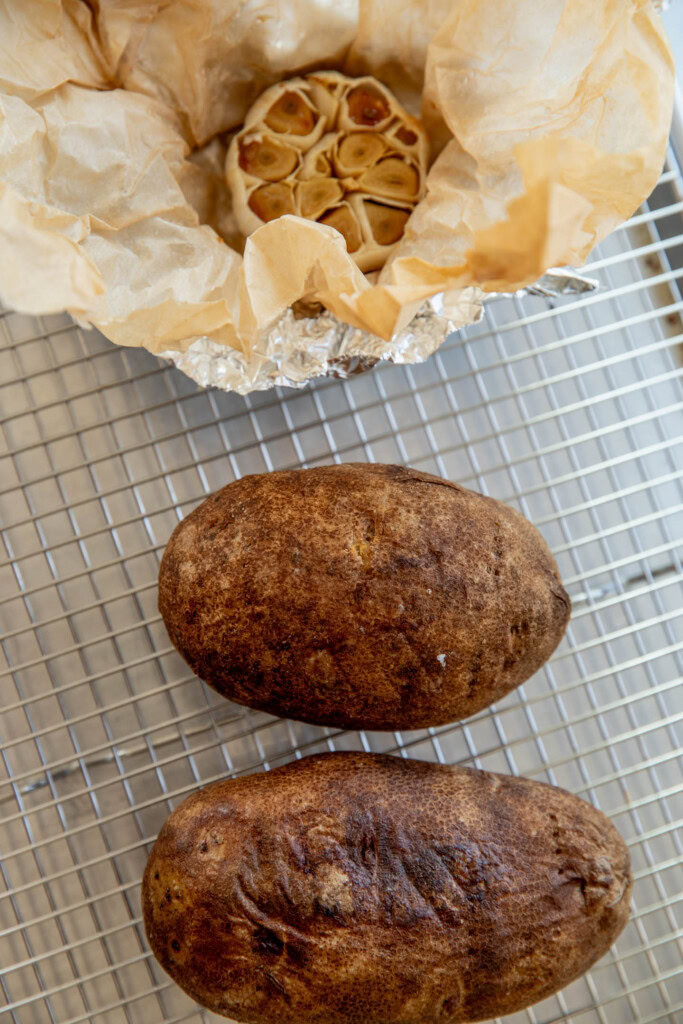 Fully baked potatoes and garlic bulb on a wire rack.