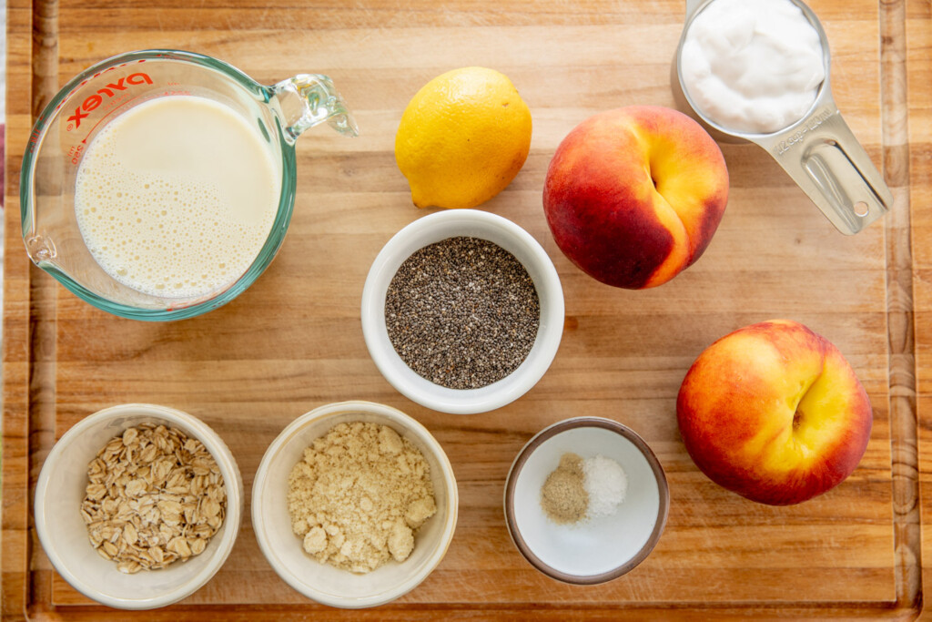 Cutting board topped with peaches, chia seeds, oats, almond flour, cardamom, milk, yogurt, and lemon on top.