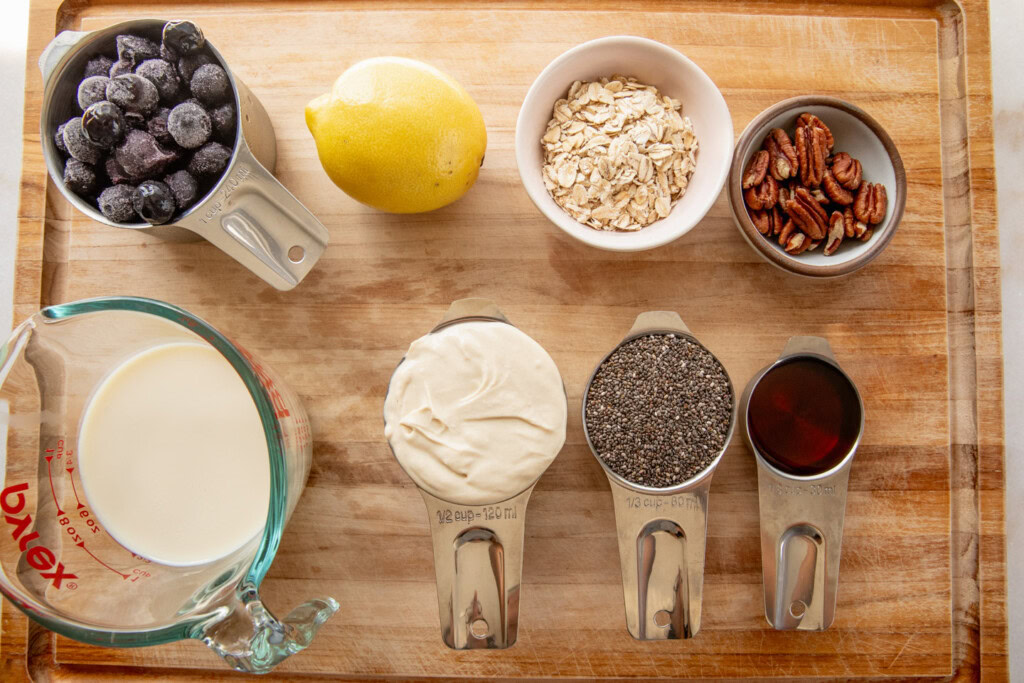 Cutting board topped with frozen blueberries, lemon, oats, pecans, chia seeds, milk, yogurt, and maple syrup.