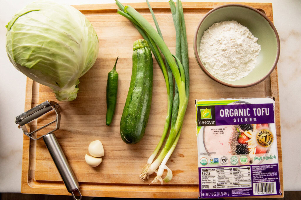 Cutting board topped with cabbage, scallions, serrano, zucchini, garlic, silken tofu, and flour.