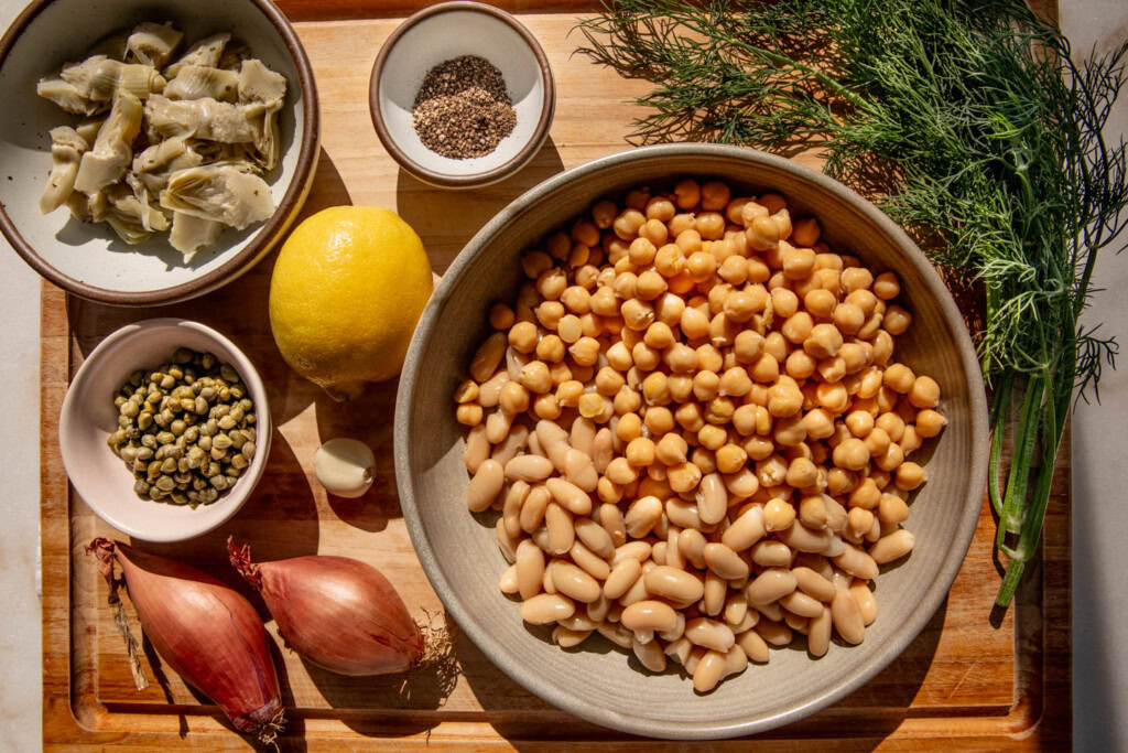 Cutting board filled with a bowl of beans, shallots, lemon, dill, artichoke hearts, and capers.