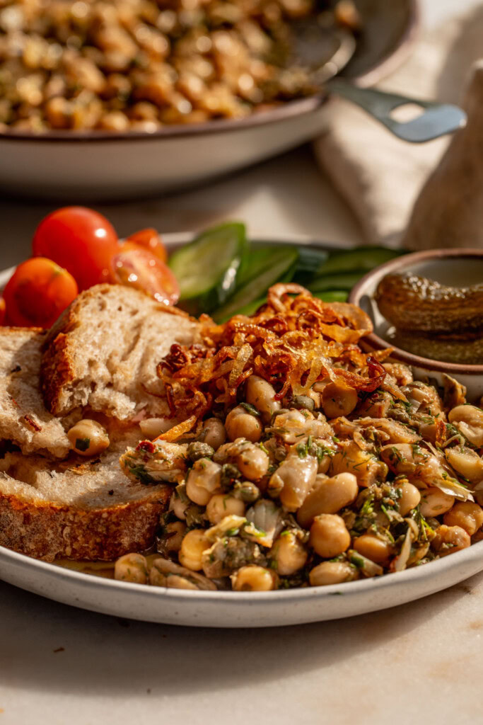 Close up of the marinated artichoke heart bean salad topped with crispy shallots, cherry tomatoes, cucumbers, bread, and gherkins.