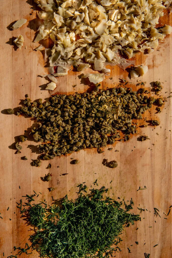 Cutting board filled with minced dill, artichoke hearts, and capers.