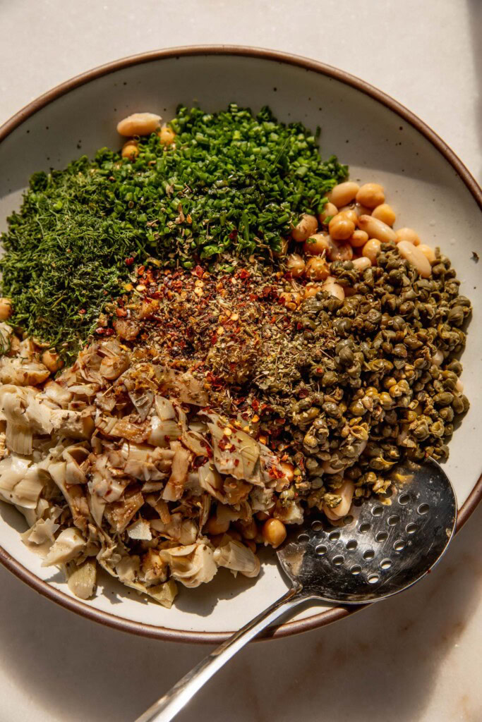 Mixing bowl filled with beans, chives, dill, capers, artichoke hearts, oregano, and garlic.