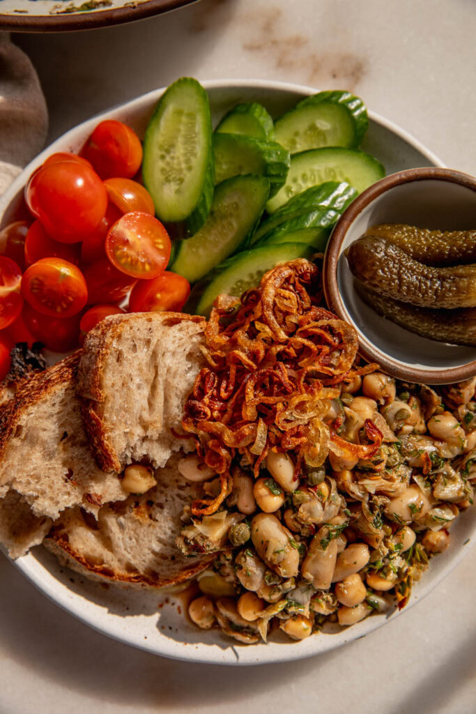 Close up of a snacking plate filled with a marinated bean salad topped with crispy shallots, torn pieces of bread, cherry tomatoes, cucumbers, and gherkins.