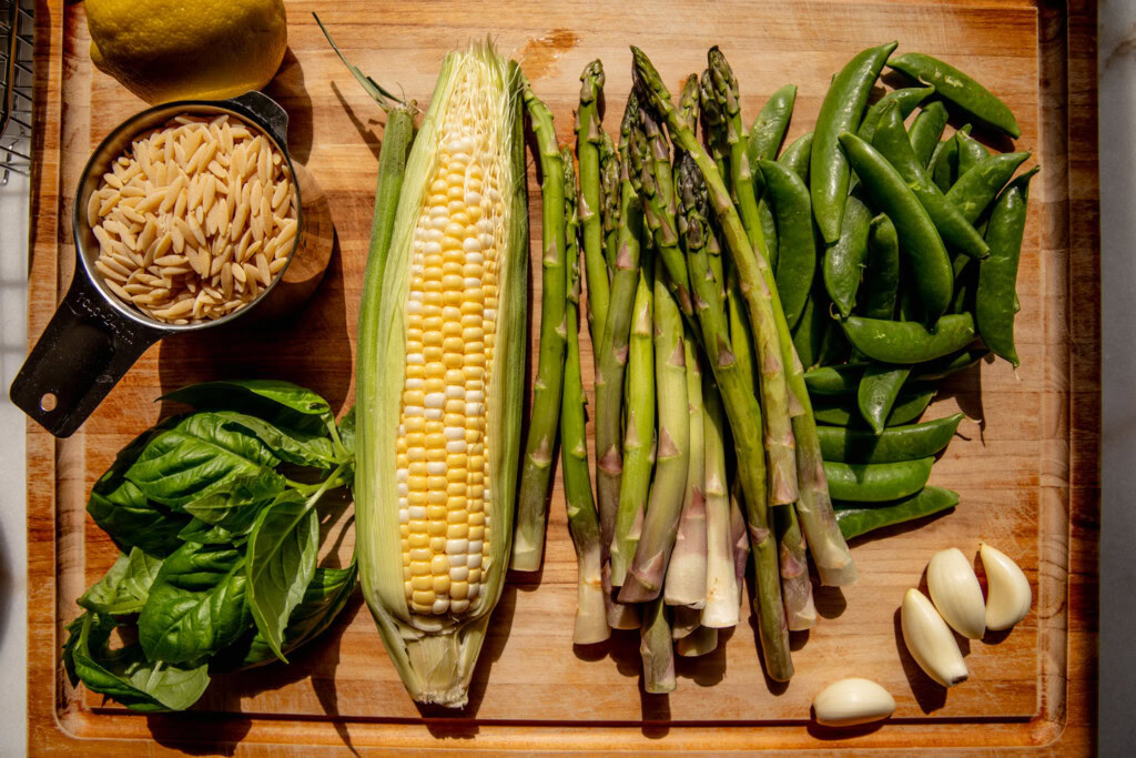 Cutting board topped with corn cobs, asparagus, snap peas, orzo, basil, shallot.
