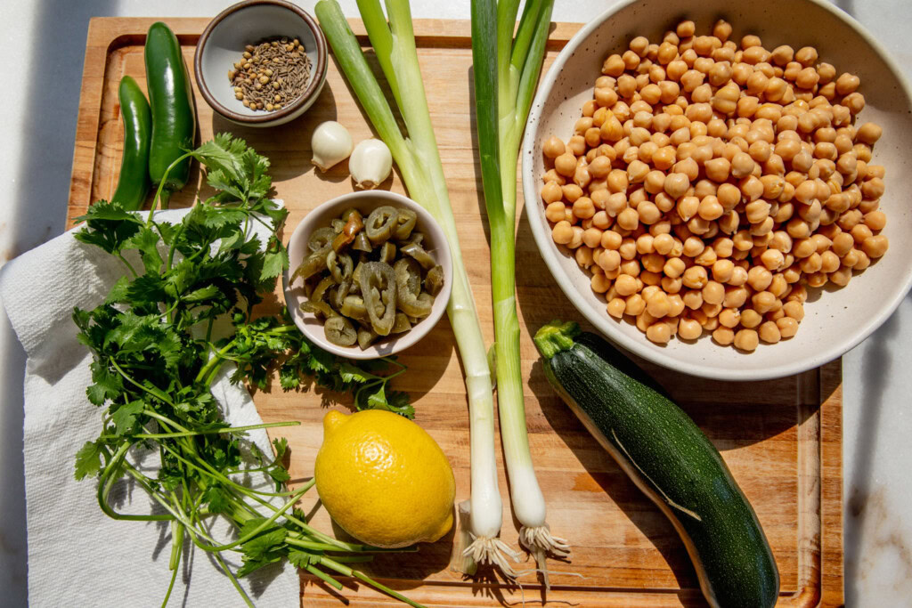Cutting board topped with a bowl of chickpeas, scallions, zucchini, pickled jalapeños, lemon, cilantro, spices, garlic and green peppers.