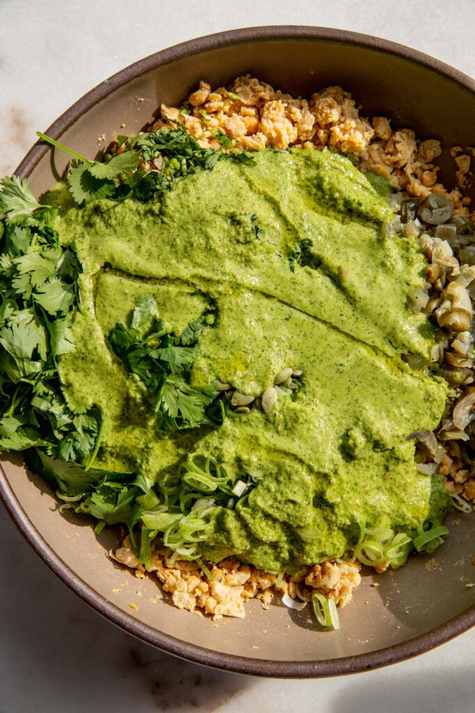 Pouring the sauce on top of the bowl of mashed chickpeas, herbs, pickled jalapeños, scallions and pepitas.
