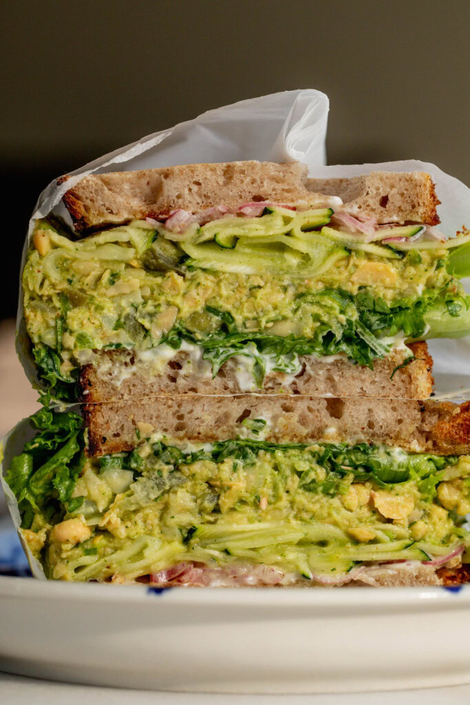 A sandwich cut in half to reveal the layers of lettuce, chickpea filling, marinated zucchini, and pickled onions.