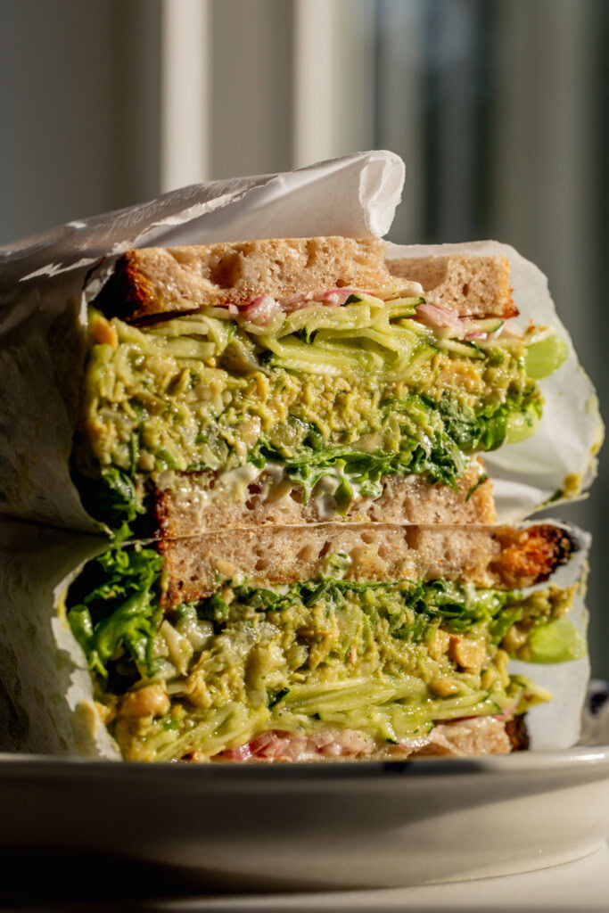 Side view of a sandwich cut in half and stacked on top of itself revealing a center layered with lettuce, chickpea filling, zucchini, and pickled onions.