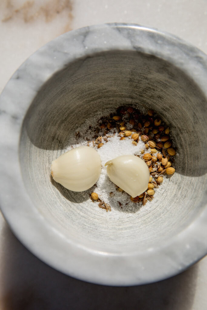 Adding the toasted seeds to a mortar and pestle with garlic cloves, and salt.