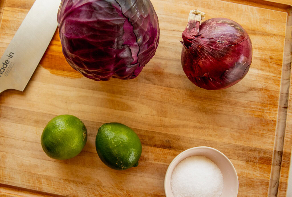 Cutting board with red cabbage, red onion, limes, and salt on the surface.