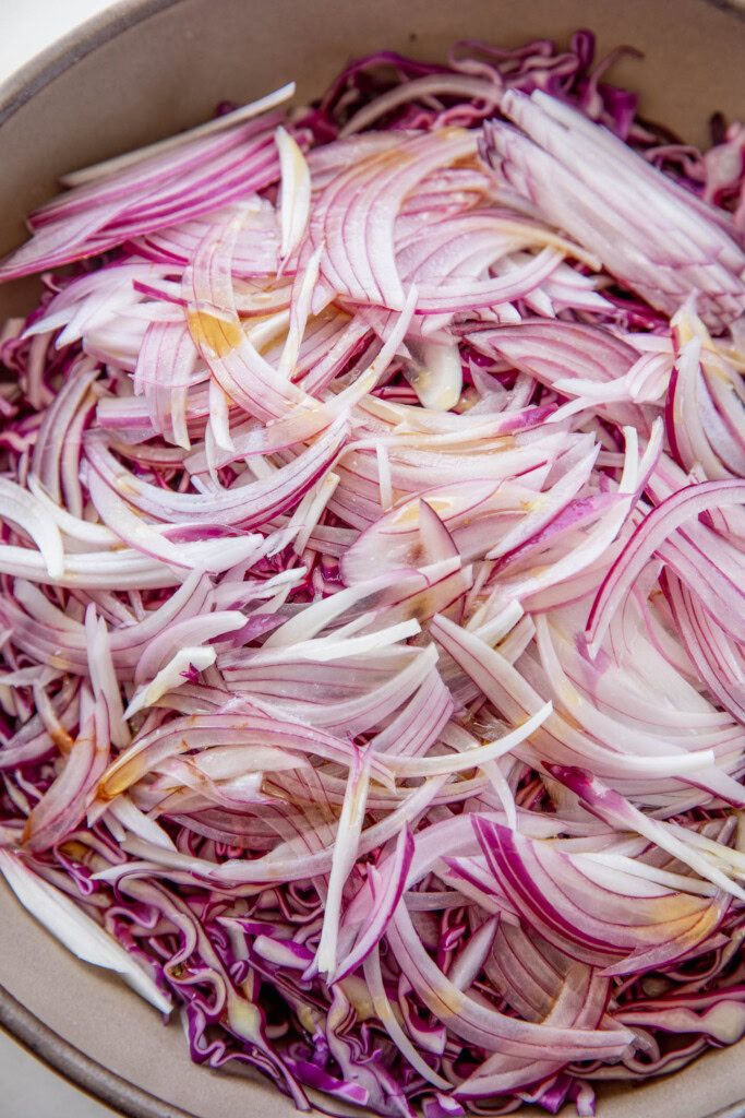 Bowl of thinly sliced cabbage, red onion, oil, maple syrup, lime juice, and salt.