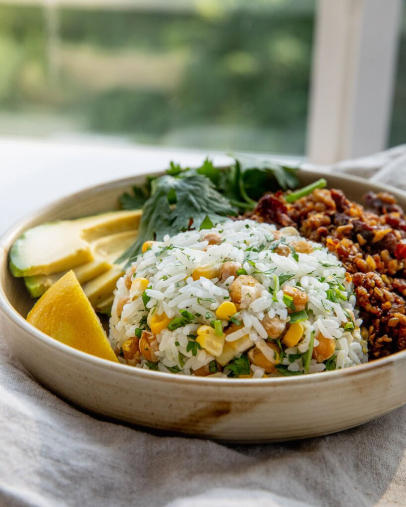 Close up of a bowl with a portion of chickpea rice in a bowl with slices of avocado and tempeh behind it.