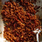 Spiced and shredded tempeh with sun-dried tomatoes in a pan being stirred together with a spoon.