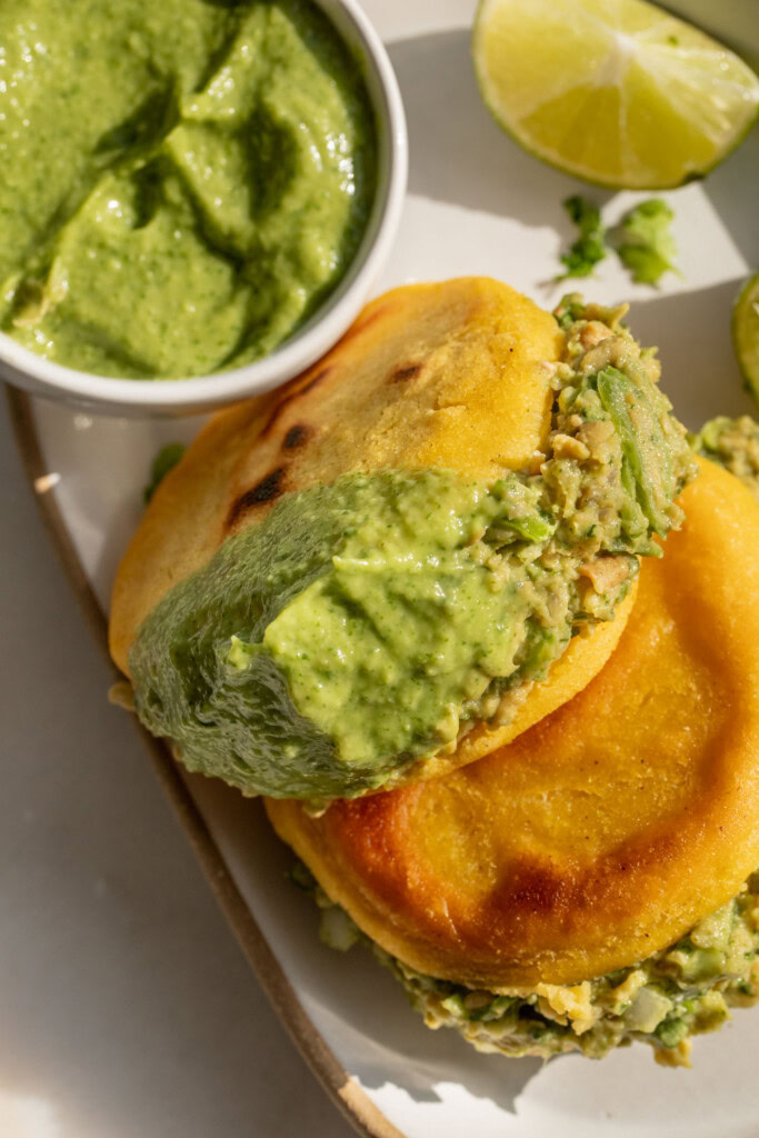 An arepa stuffed with tofu salad after being dipped in extra guasacaca on a plate.