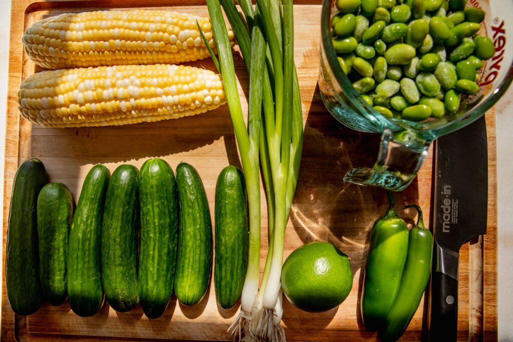 Cutting board with Persian cucumbers, corn on the cob, scallions, edamame, lime, and serrano peppers.
