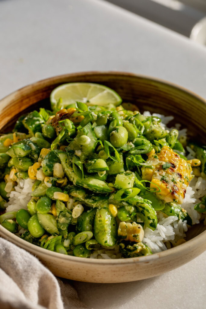Side view of a bowl of rice topped with corn and edamame salad with a wedge of lime tucked into the back.