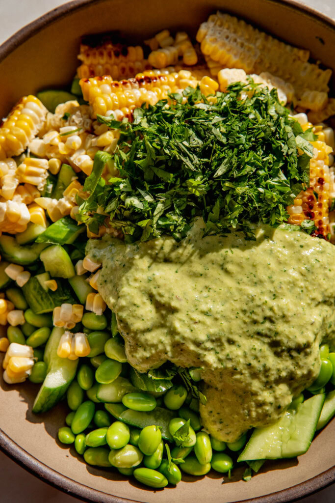 Mixing bowl with corn, smashed cucumber, cilantro, edamame with creamy scallion dressing pour on top.