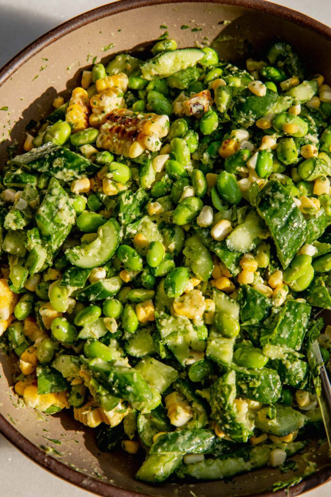 Top down shot of a mixing bowl mixed with corn, cucumbers, edamame, and scallion dressing.