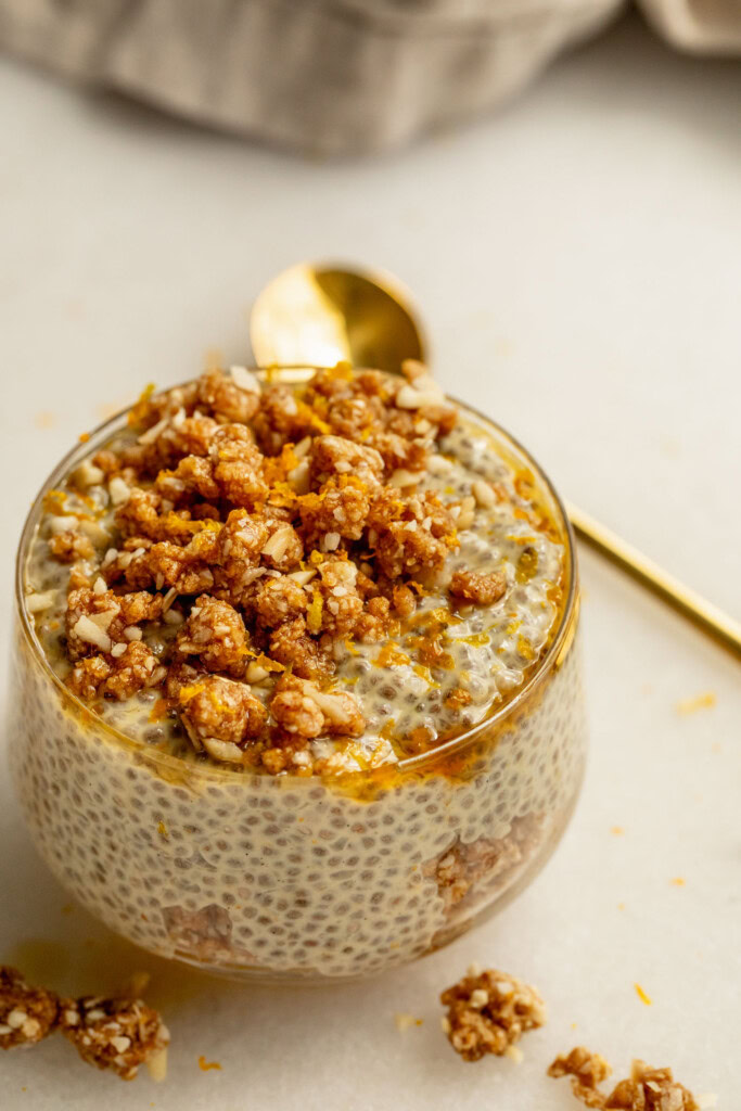 Close up of a jar of chia pudding layered with crumble, a gold spoon set behind it.