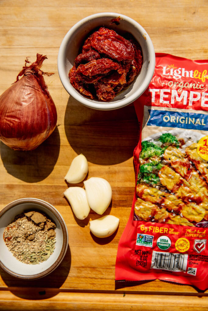 Cutting board filling with shallots, sun-dried tomatoes, spices, garlic, and tempeh.