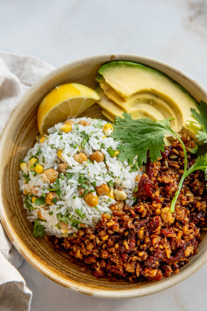 Close up of a bowl filled with rice mixed with chickpeas, shredded tempeh, avocado slices, and a wedge of lemon.