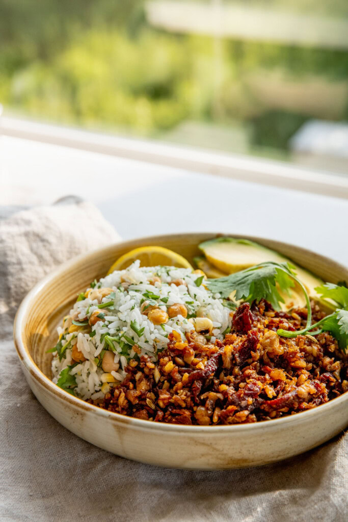Side view of a bowl filled with rice mixed with chickpeas, shredded tempeh, avocado slices, and a wedge of lemon.
