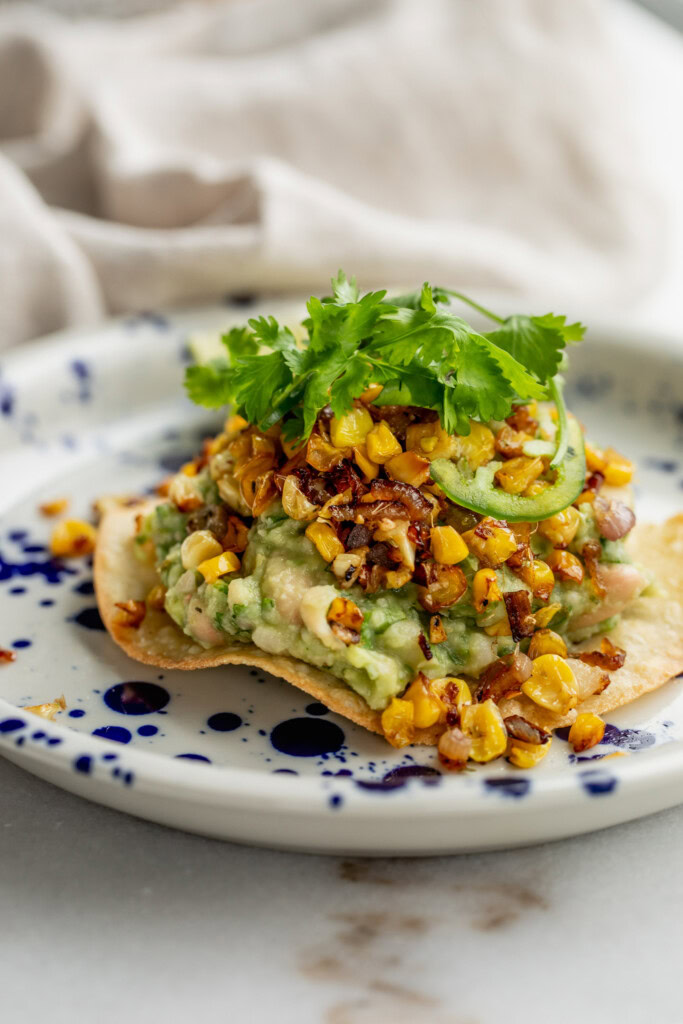Tostada on a speckled plate topped with mashed avocado and bean mash, crispy corn, and cilantro.