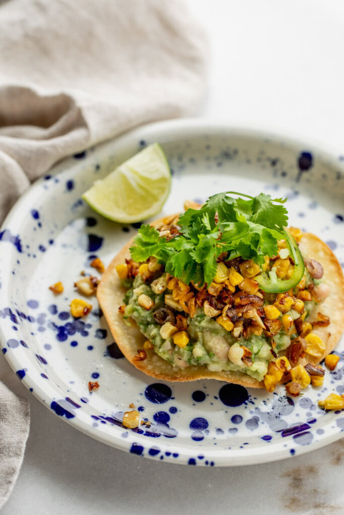 Upward view of a tostada topped with white bean avocado mash, crispy corn, and cilantro.